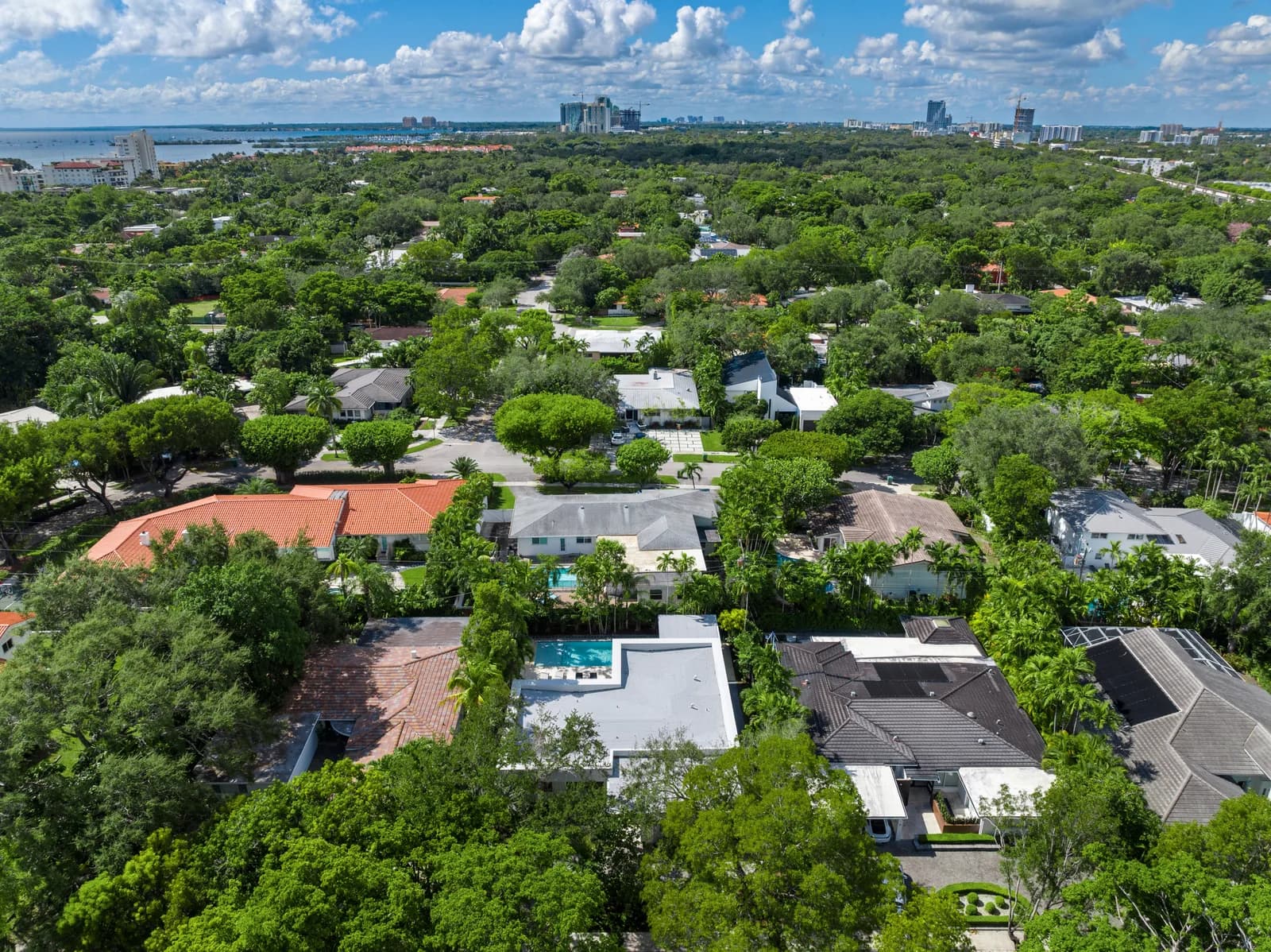 Aerial view over Coconut Grove rooftops, canopy and adjacent lots with the Miami skyline and Biscayne Bay in the distance, showing how one property sits against the zoning and setbacks of the lots around it
