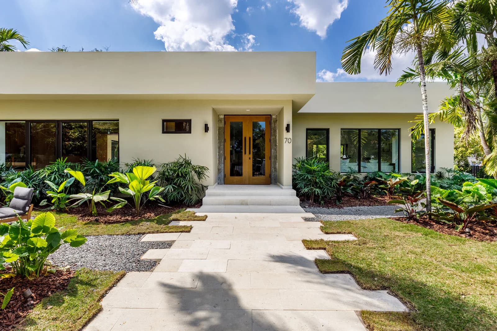 The stucco and warm wood entry facade of Luxom's Samana Residence in Coconut Grove, with a travertine walkway and tropical landscaping either side of the front door